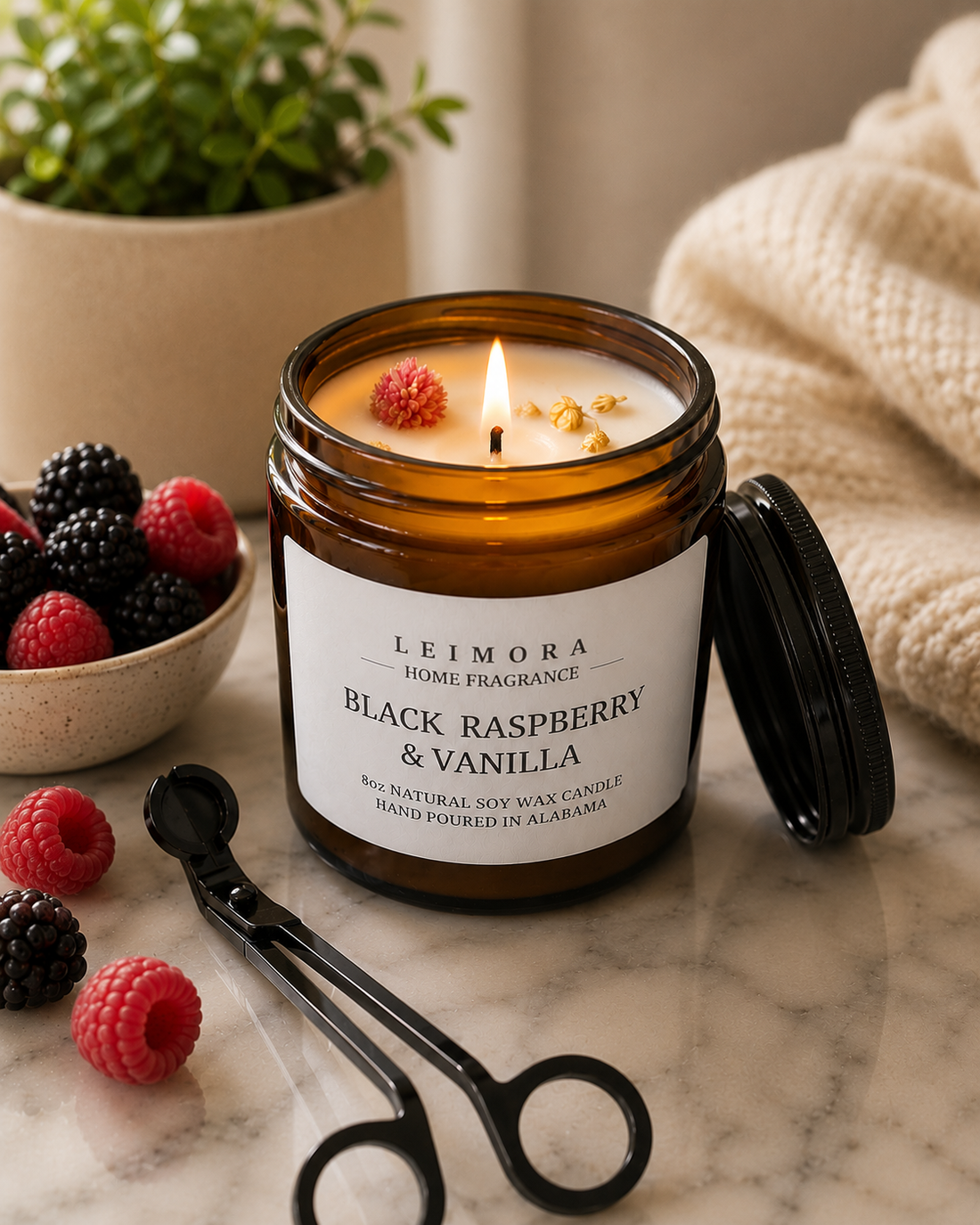 Lit Black Raspberry & Vanilla soy candle from Leimora Home Fragrance in an amber glass jar, featuring a full melt pool and decorative wax accents, styled on a marble countertop with fresh berries, a wick trimmer, a cozy knit blanket, and a potted plant.